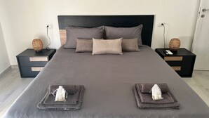 Ein ordentlich bezogenes Bett mit dunkelgrauer Tagesdecke und mehreren Kissen, flankiert von zwei schwarzen Nachttischen mit gewobenen Lampen und einer kleinen Schublade.