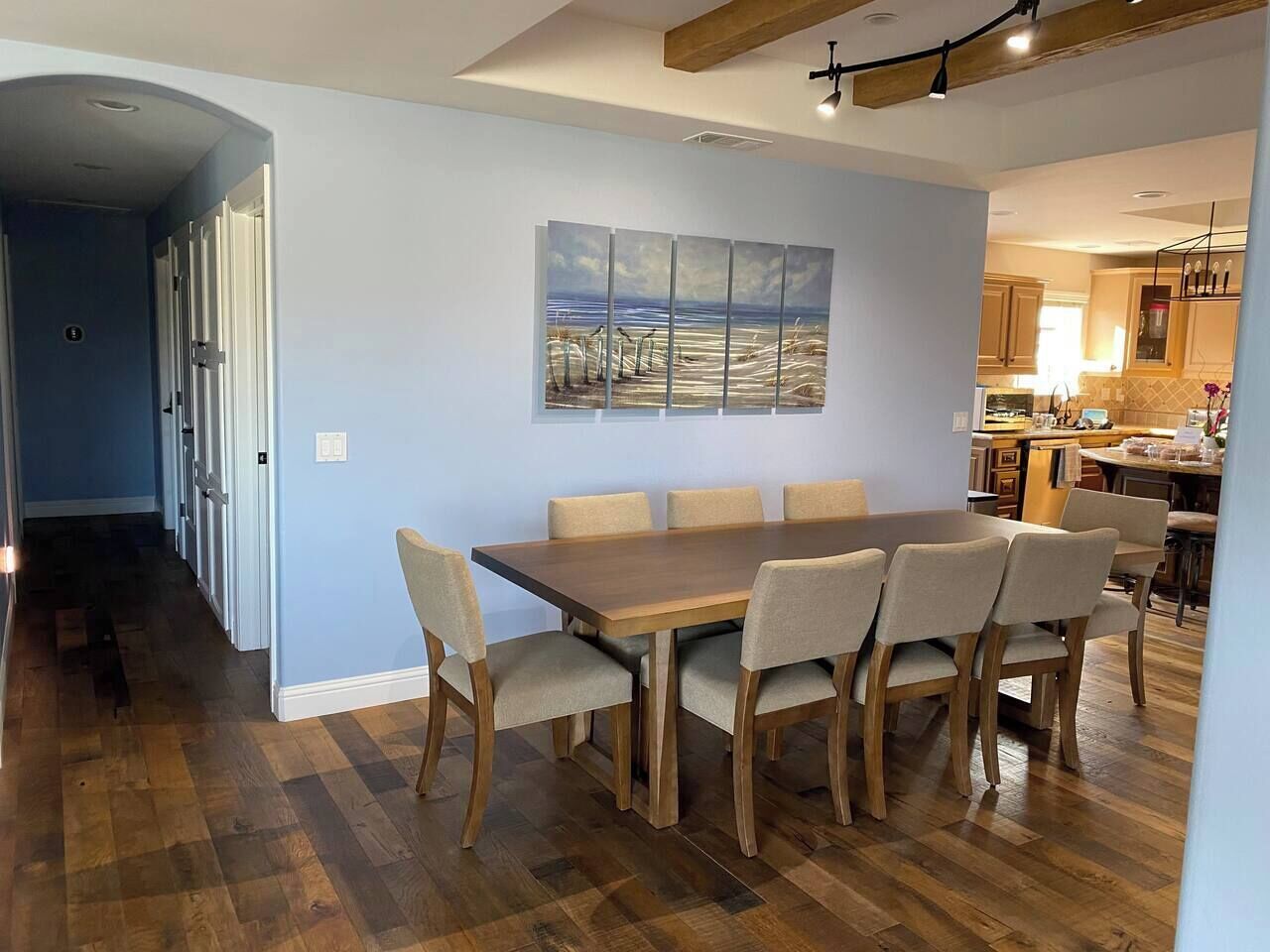 A dining area with a wooden table and chairs, a painting on the wall, and a kitchen in the background.