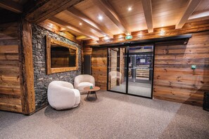 A log cabin interior with stone walls, wooden ceiling beams, a stone fireplace, two white armchairs, and a small round table. - Hôtel Emeraude (Landry)