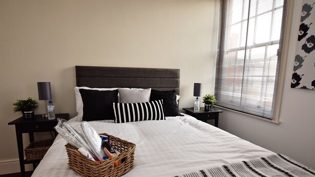 A neatly made bed with a striped blanket, a wicker basket with items, and a window with blinds. - Derby 2 Bed House - University & City (Derby)