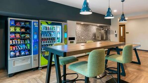 Un hall d’hôtel moderne, doté de distributeurs automatiques, d’une table à manger en bois et de chaises recouvertes d’un tissu vert.