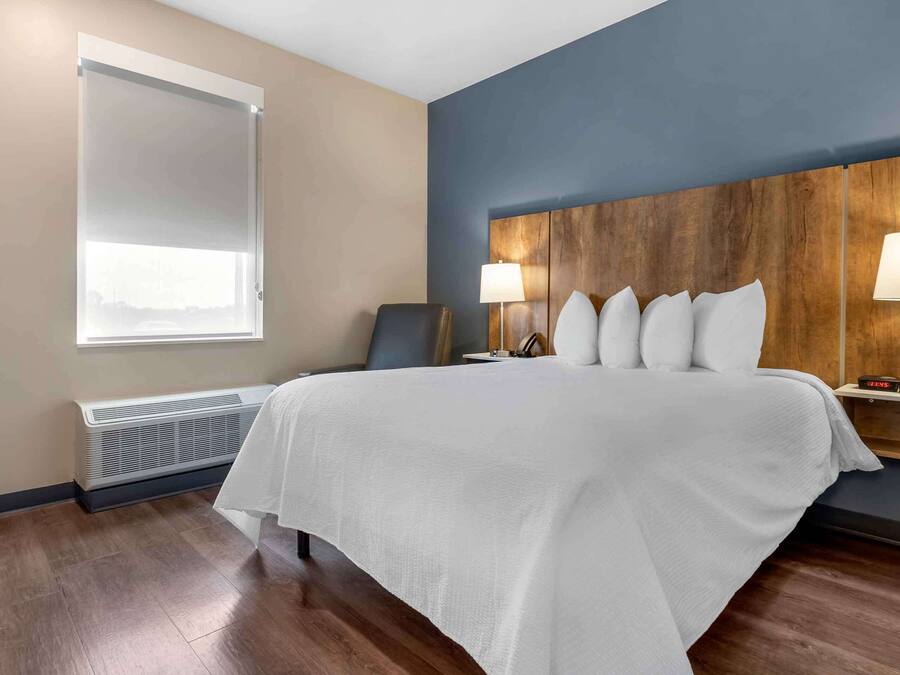A hotel room with a large bed, a wooden headboard, two bedside tables with lamps, a window with blinds, and a wall-mounted heater.