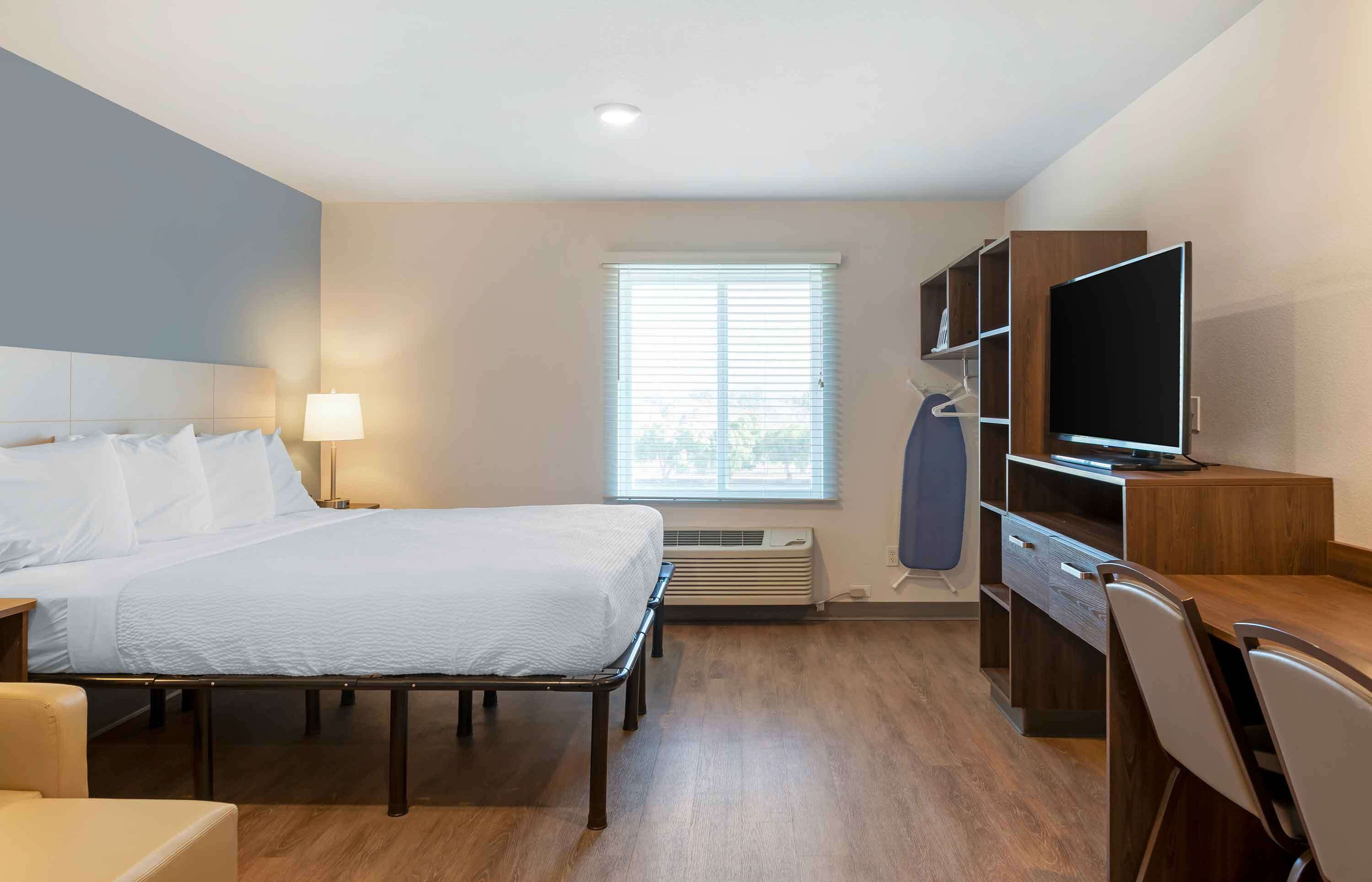 A hotel room with a bed, bedside table, lamp, TV, desk, chair, and a window with blinds.