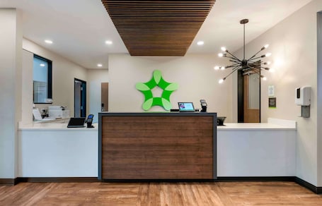 A reception desk with a wooden panel, a green abstract wall art, a digital screen, and a modern chandelier.. Extended Stay America Premier Suites - Colorado Springs - South