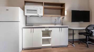 A compact kitchen with a microwave, a refrigerator, and a sink. There is a wooden shelf above the microwave and a flat-screen TV mounted on the wall.