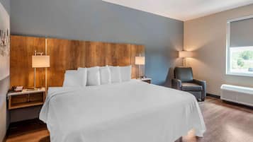 A modern hotel room with a large bed, wooden headboard, two bedside tables with lamps, a chair, and a window with a view.