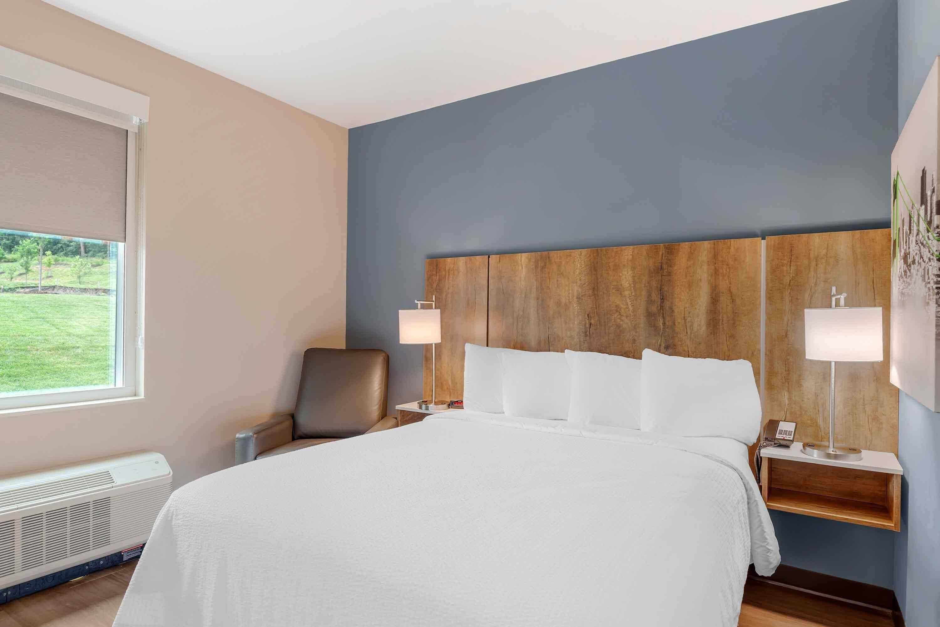 Une chambre d’hôtel avec un grand lit, deux tables de chevet avec des lampes, une chaise, un radiateur et une fenêtre donnant sur un paysage verdoyant.