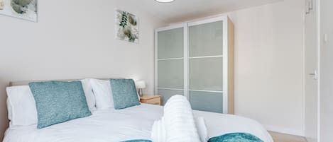 A neatly made bed with a teal blanket and white pillows, a bedside lamp, and a wall with a framed botanical print.