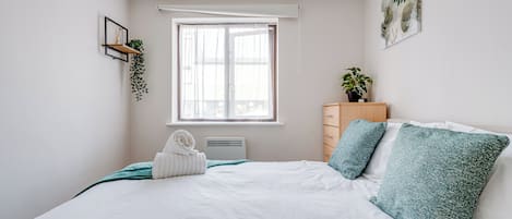 A bedroom with a bed, a window, a wall-mounted shelf with a plant, a bedside table with a plant, and a wall art piece.