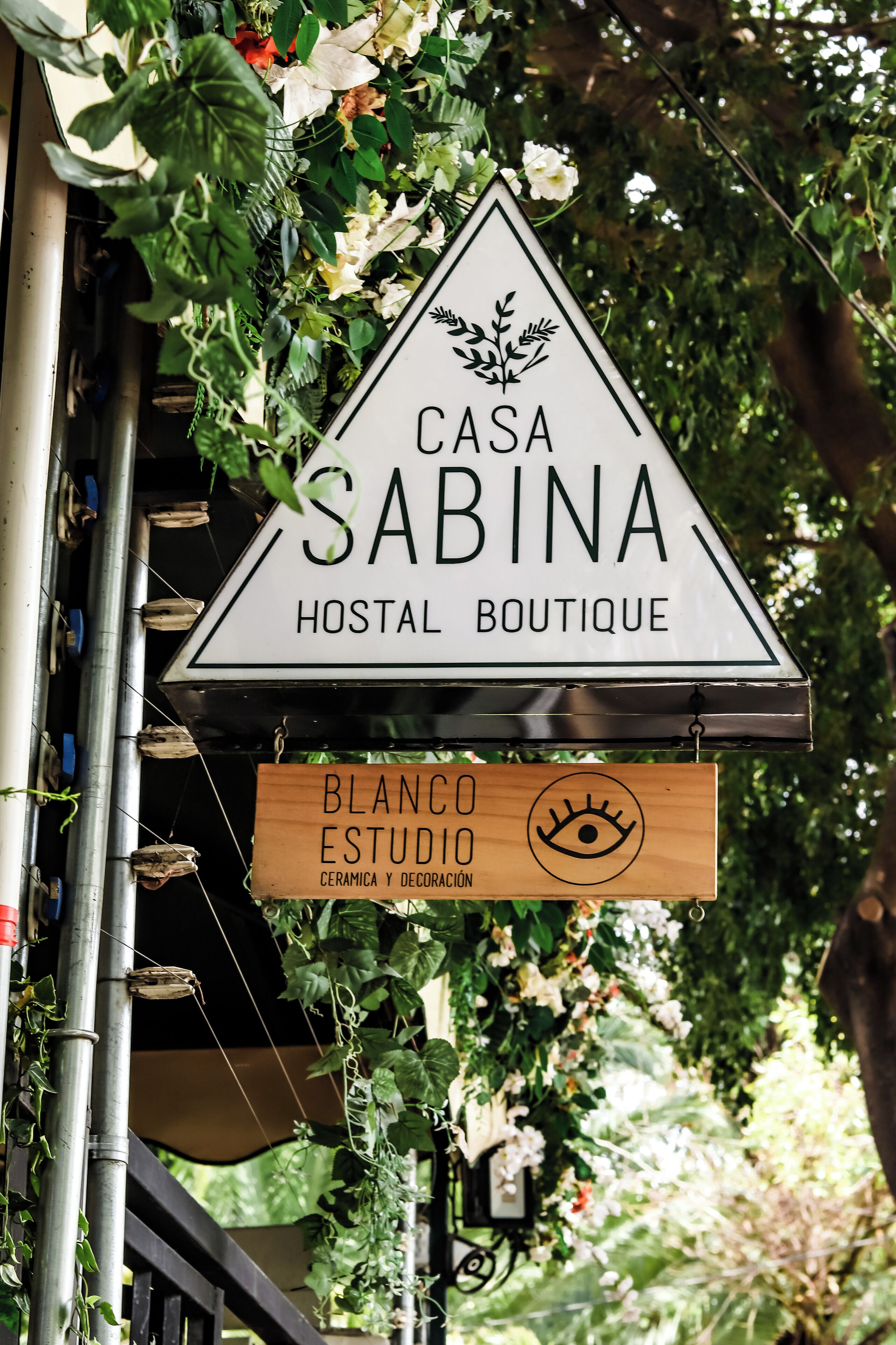 A sign for 'Casa Sabina Hostel Boutique' with a triangular design and a logo above the name. Below it, a wooden sign reads 'Blanco Estudio Cerámica y Decoración' with an eye symbol.