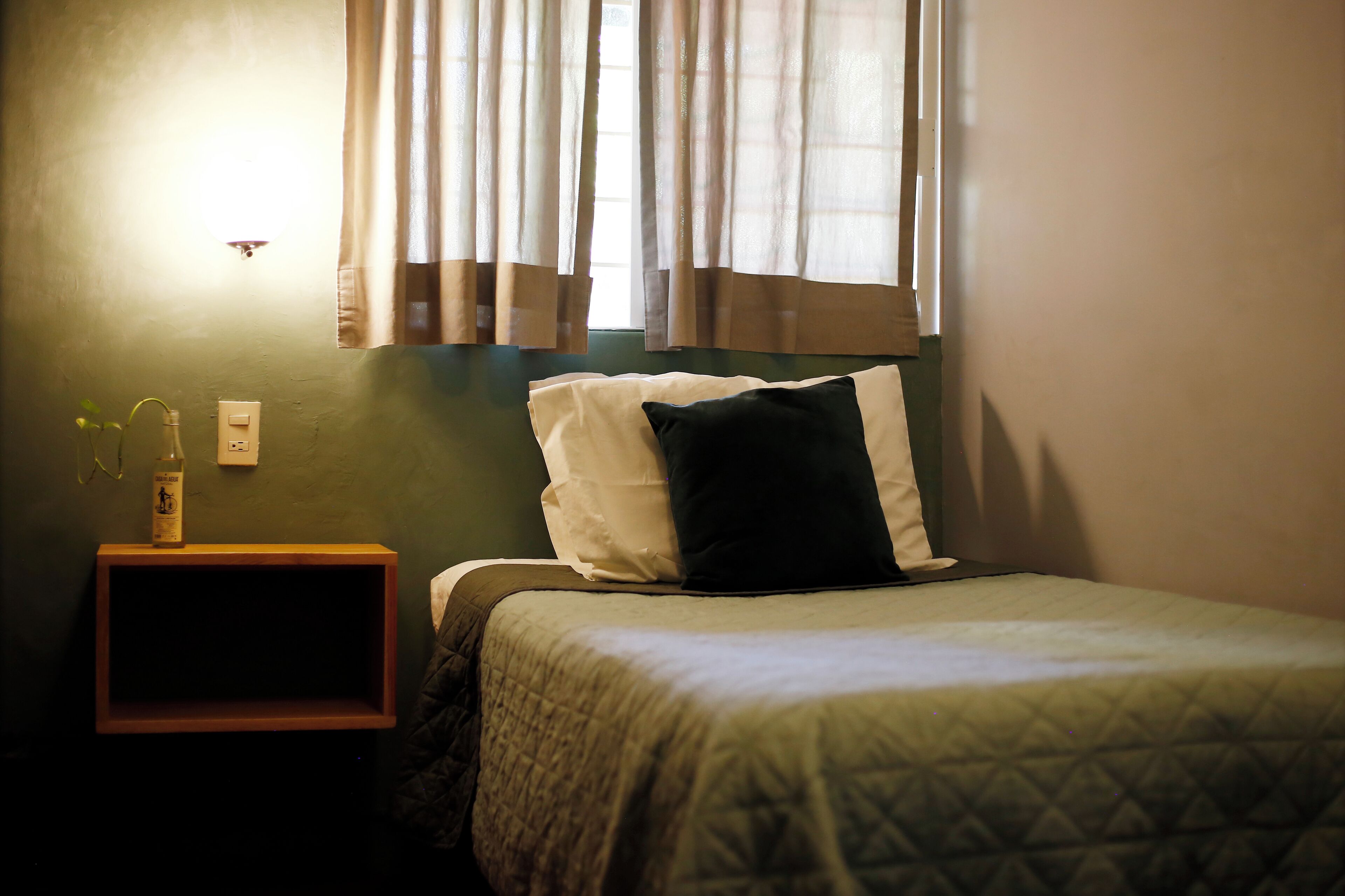A small, neatly made bed with a quilted bedspread and two pillows, a bedside table with a bottle and glasses, and a window with curtains.