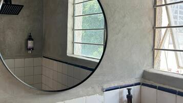 A bathroom with a round mirror, a window, and a wooden shelf with a bottle.