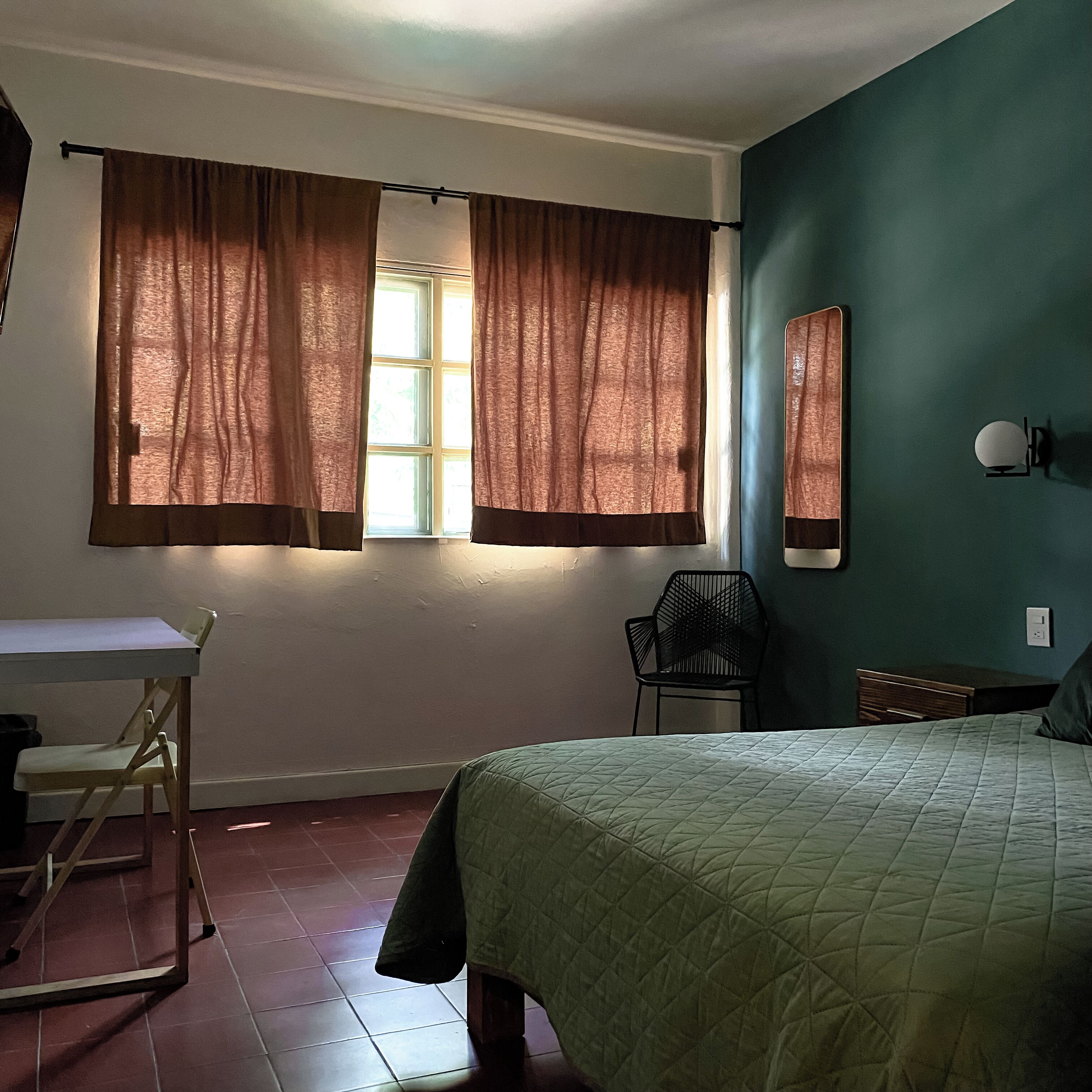 Une pièce comprenant un lit, un bureau, une chaise et une fenêtre avec des rideaux.