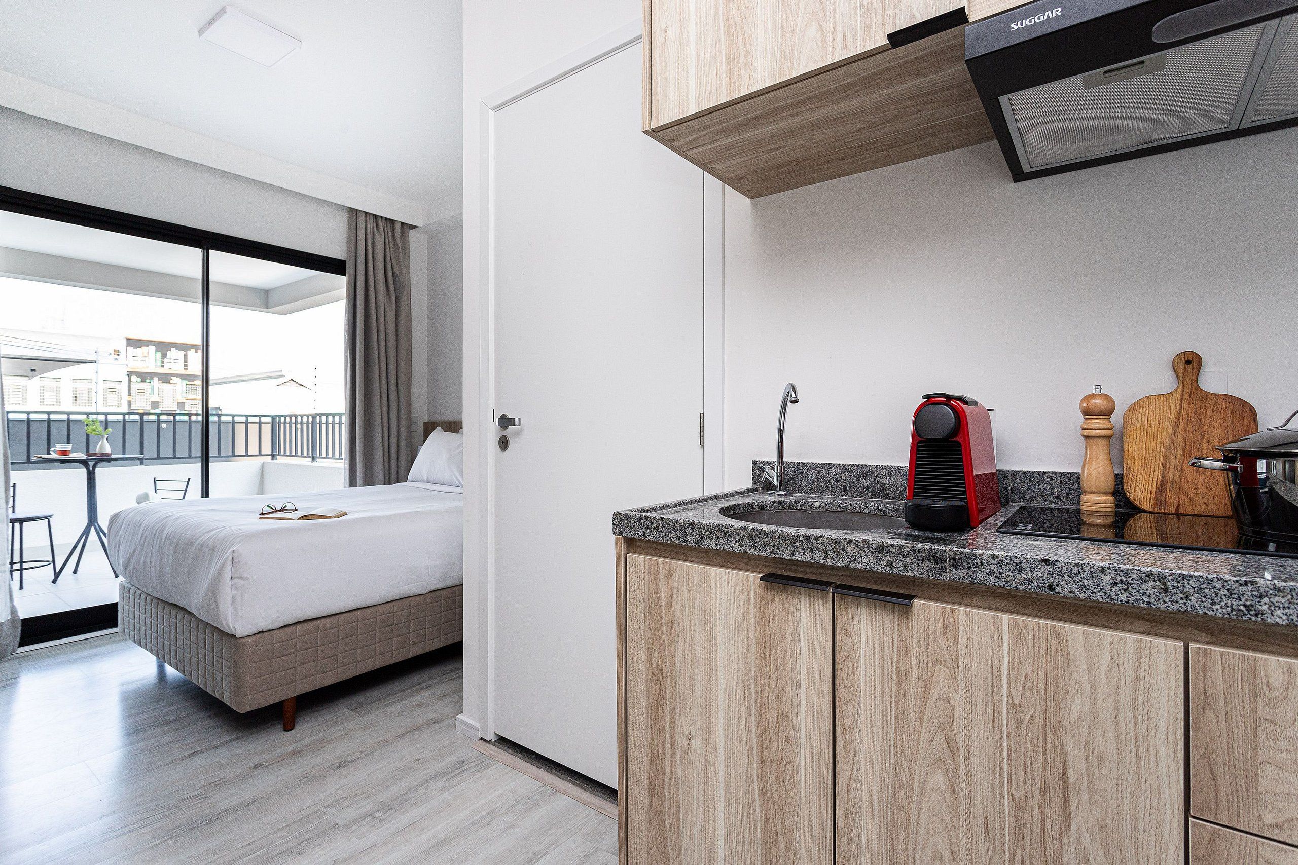 A modern hotel room with a bed, a small kitchenette, and a balcony with a view.