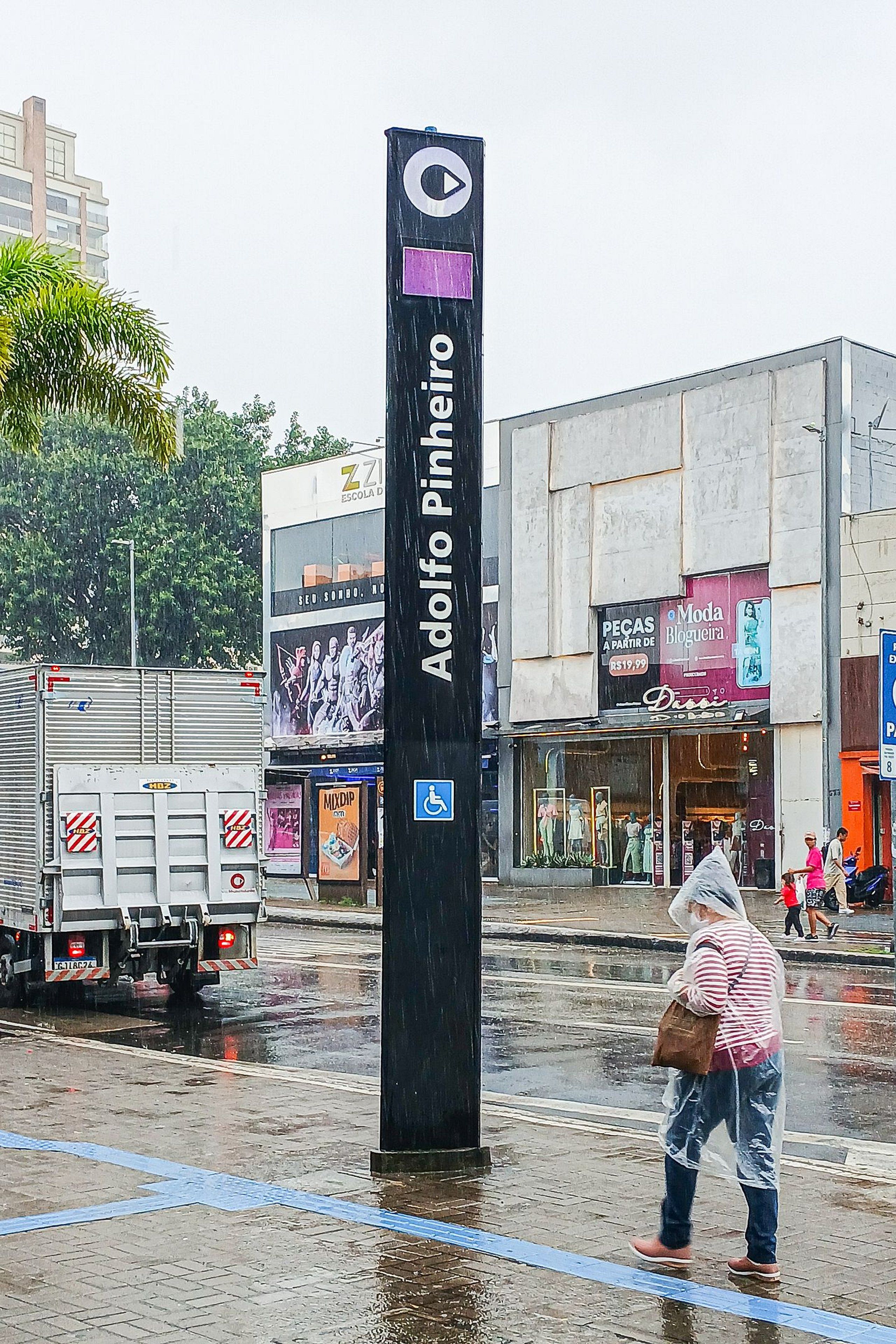 A wet urban street scene with a prominent black signpost reading 'Ado Pinho'.
