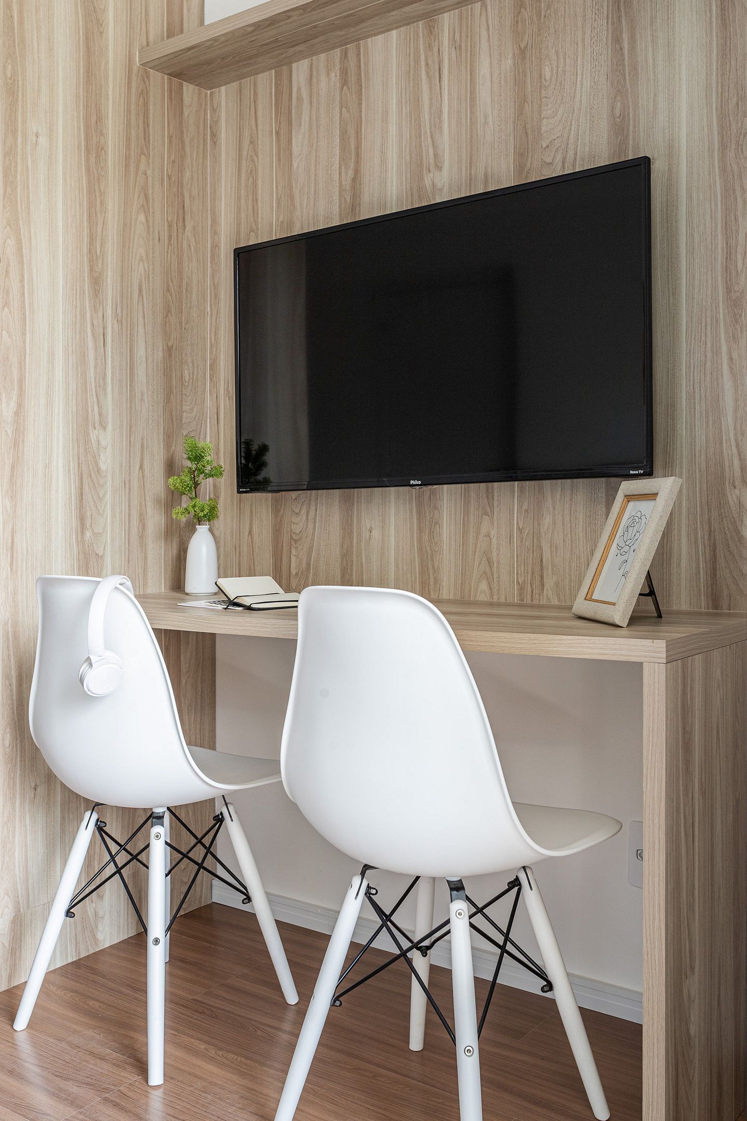 Ein modernes Zimmer mit einer Holzvertäfelung, einem wandmontierten Flachbildfernseher, zwei weißen Stühlen, einem kleinen weißen Tisch mit einer Vase und einem Bilderrahmen sowie einem weißen Schreibtisch.