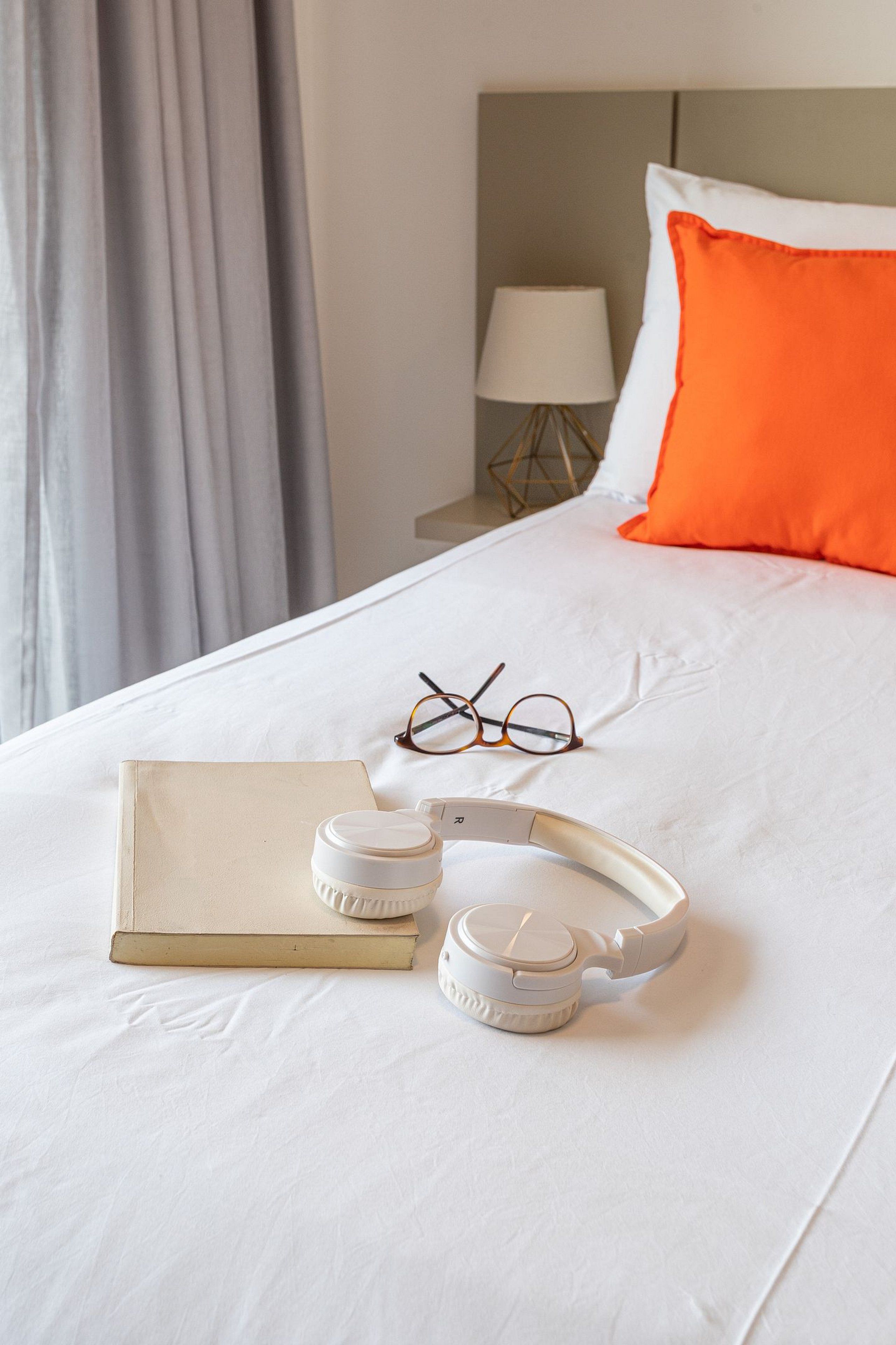 Una cama bien hecha con un edredón blanco, un par de gafas, un libro y un altavoz portátil blanco.