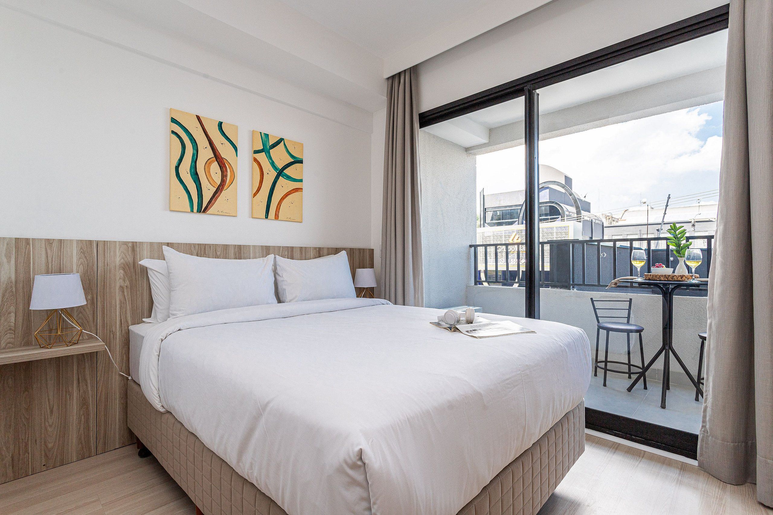 Quarto de hotel com uma cama grande, uma escrivaninha com cadeira e uma varanda com mesa e cadeiras.