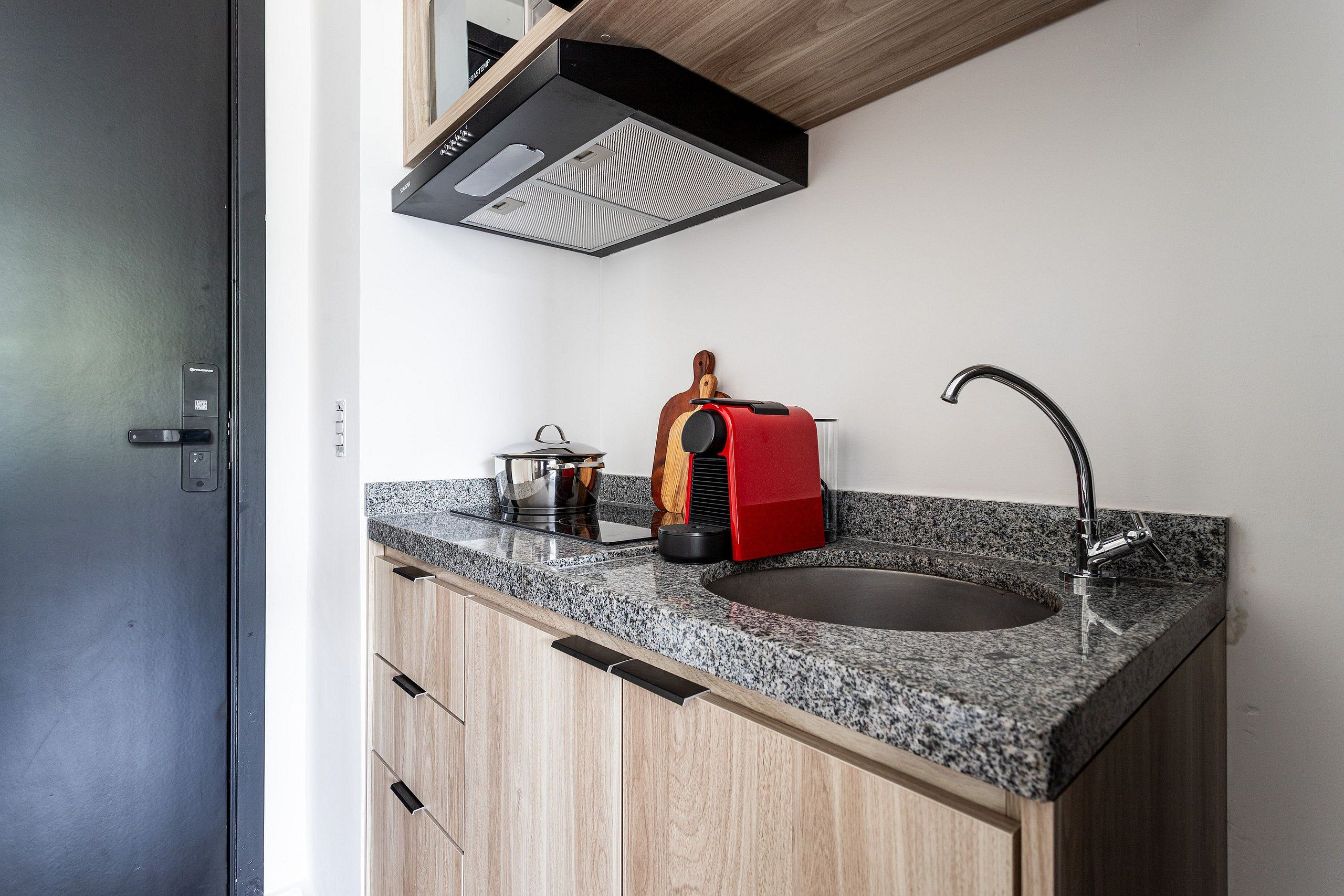 Cozinha moderna com bancada de granito, pia de aço inoxidável, cafeteira vermelha e armário de madeira.