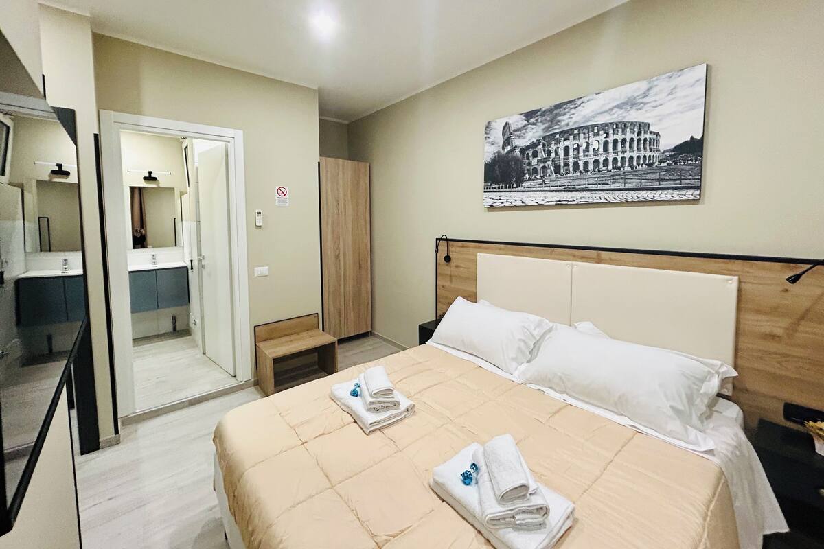 A hotel room with a bed, a bathroom with a mirror, a wardrobe, and a picture of a historical building on the wall.