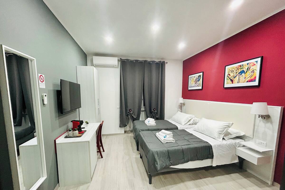 A modern hotel room with a grey and red color scheme, a flat-screen TV, a desk with a coffee machine, two beds, and artwork on the walls.