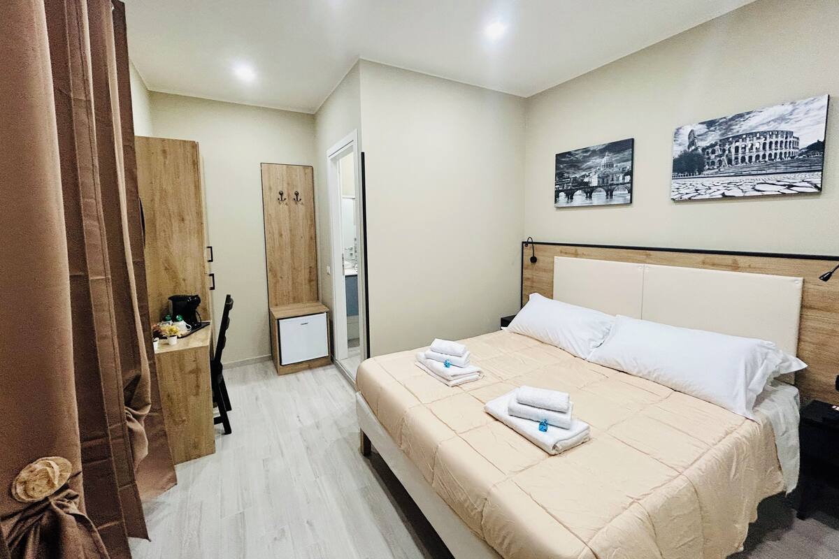 A hotel room with a bed, a wardrobe, a small desk, and a nightstand. There are two paintings on the wall.