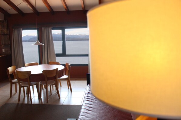 A dining area with a round table and chairs, a sofa, and a view of a body of water with mountains in the distance. - Belgrano Decks II Rental Apartments (San Carlos de Bariloche)