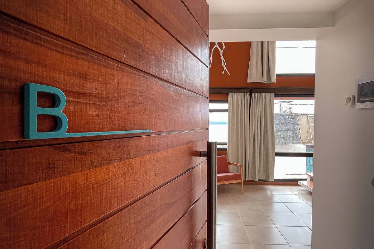 Un mur en bois sur lequel est fixée une lettre « B », une porte coulissante et une vue sur une pièce avec une fenêtre et des rideaux.