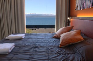 Una cama con vista a un lago y montañas, un cabecero y una mesita de noche con una lámpara.