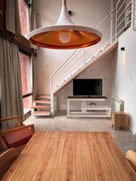 Una sala moderna con una mesa de comedor de madera, un televisor sobre un soporte blanco, una escalera con barandilla y un ventanal con cortinas.