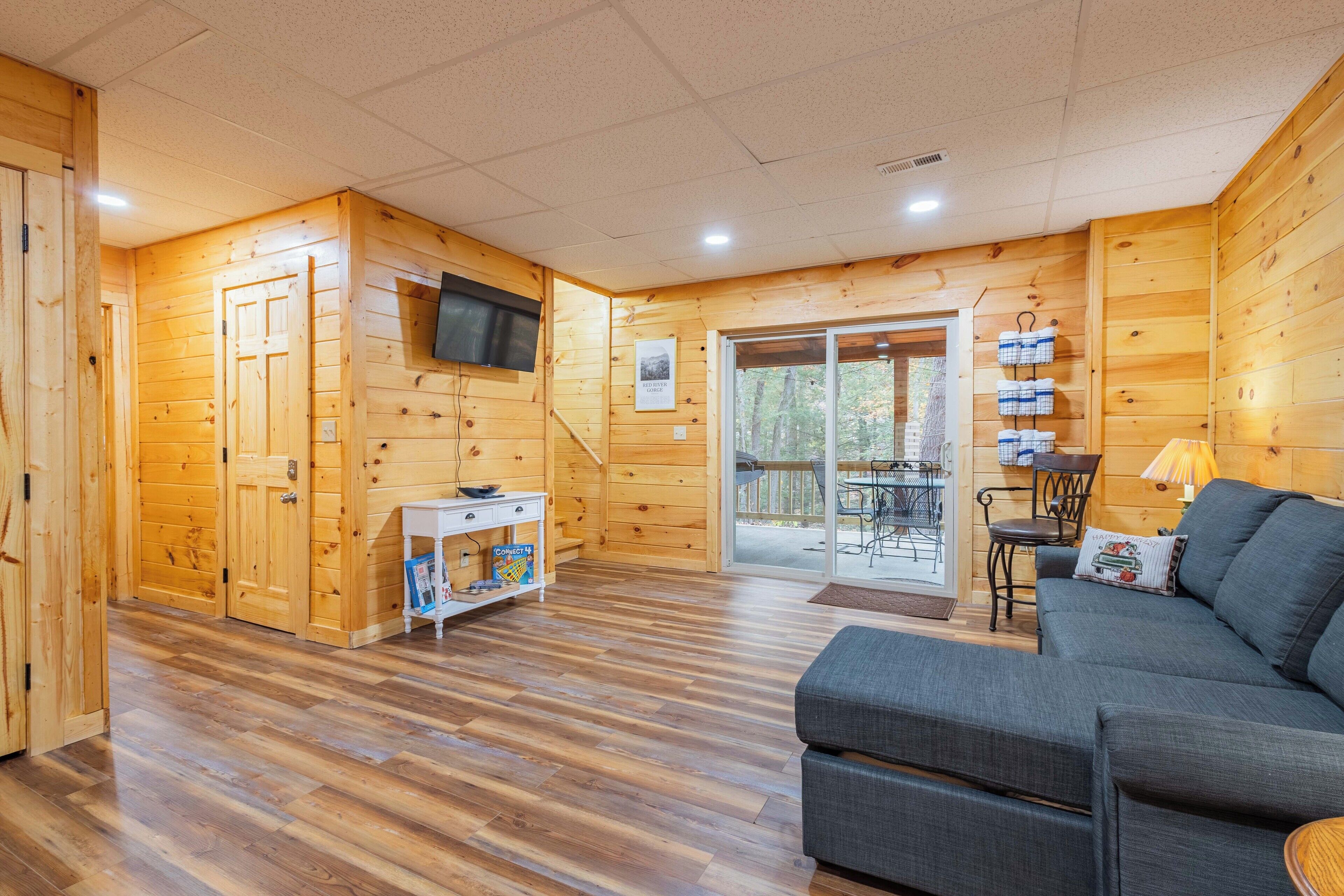 A wooden cabin interior with a sofa, a TV mounted on the wall, and a sliding glass door leading to an outdoor deck.