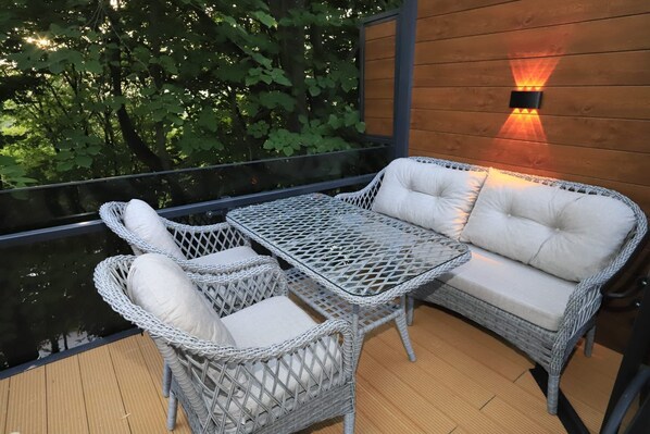 Un área de estar exterior de mimbre con sillas acolchadas y una mesa, una terraza de madera y una lámpara fijada a la pared.