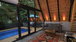 Una stanza spaziosa con una grande finestra vista piscina, un'accogliente area salotto con divano e poltrone, e un soffitto in legno.