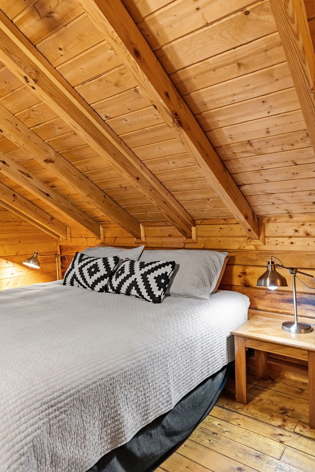 A cozy wooden cabin room with a bed, pillows, a nightstand, and a lamp.