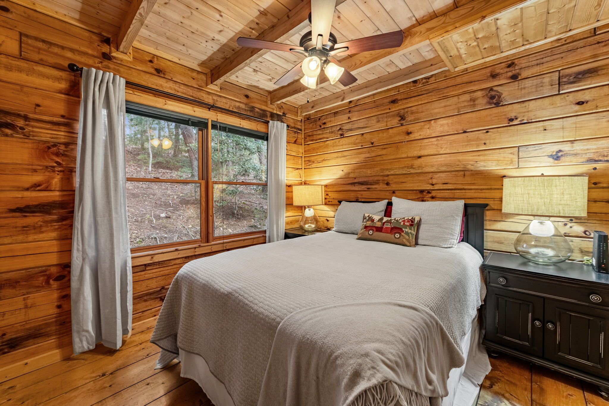 Quarto de cabana de madeira com cama, ventilador de teto e janela com cortinas.