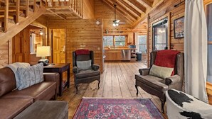 A cozy log cabin interior with a staircase, wooden walls, a kitchen area, and a living room with leather sofas and a patterned rug.