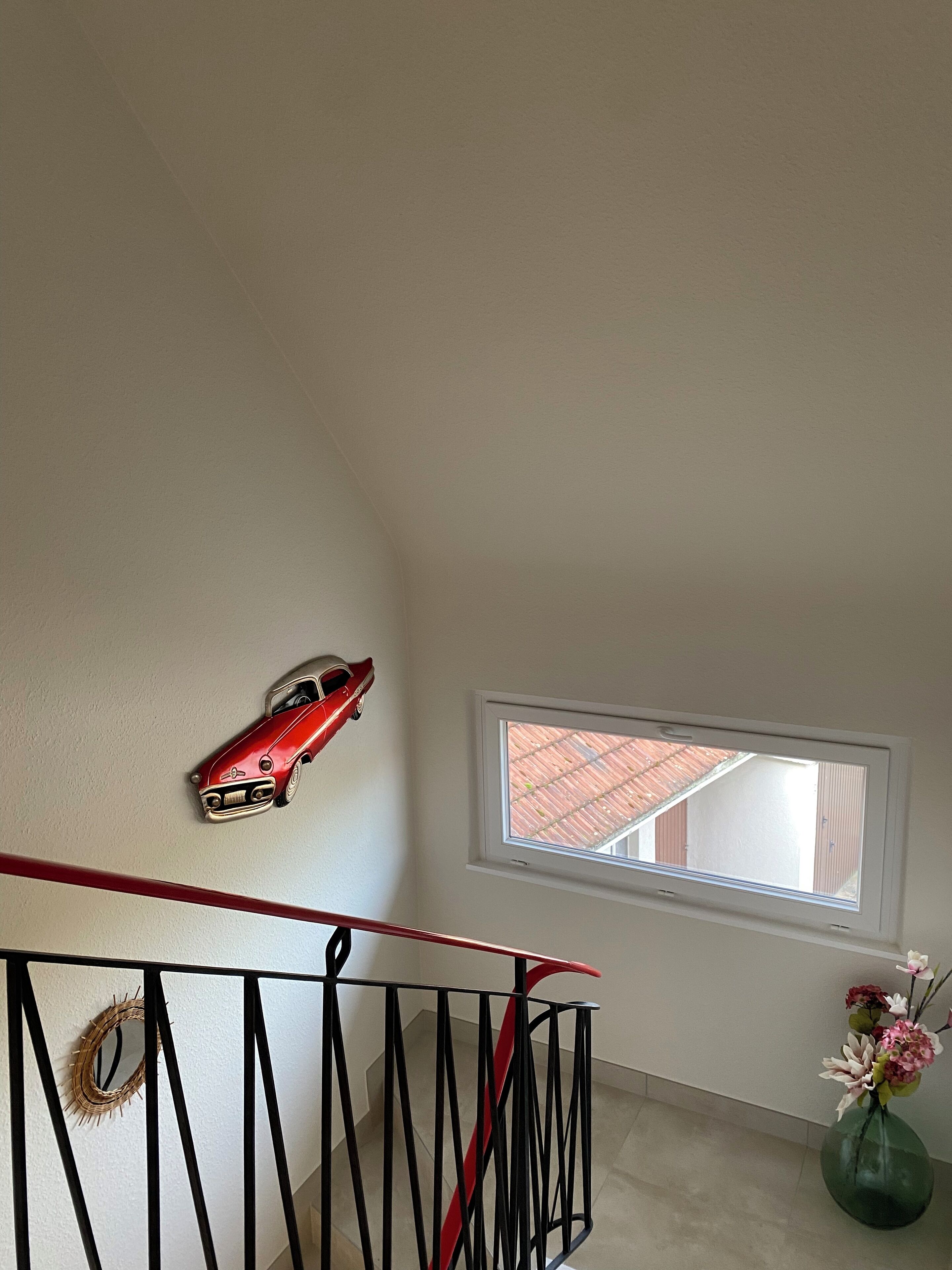 Eine Treppe mit roter Geländer, eine an der Wand befestigte Auto-Dekoration, ein Fenster mit Blick auf ein Gebäude mit rotem Dach und eine Vase mit Blumen auf dem Boden.