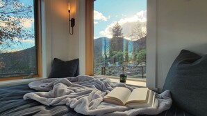 Una cama con colcha a rayas, una manta y un libro abierto. Hay una ventana con vista a árboles y colinas.