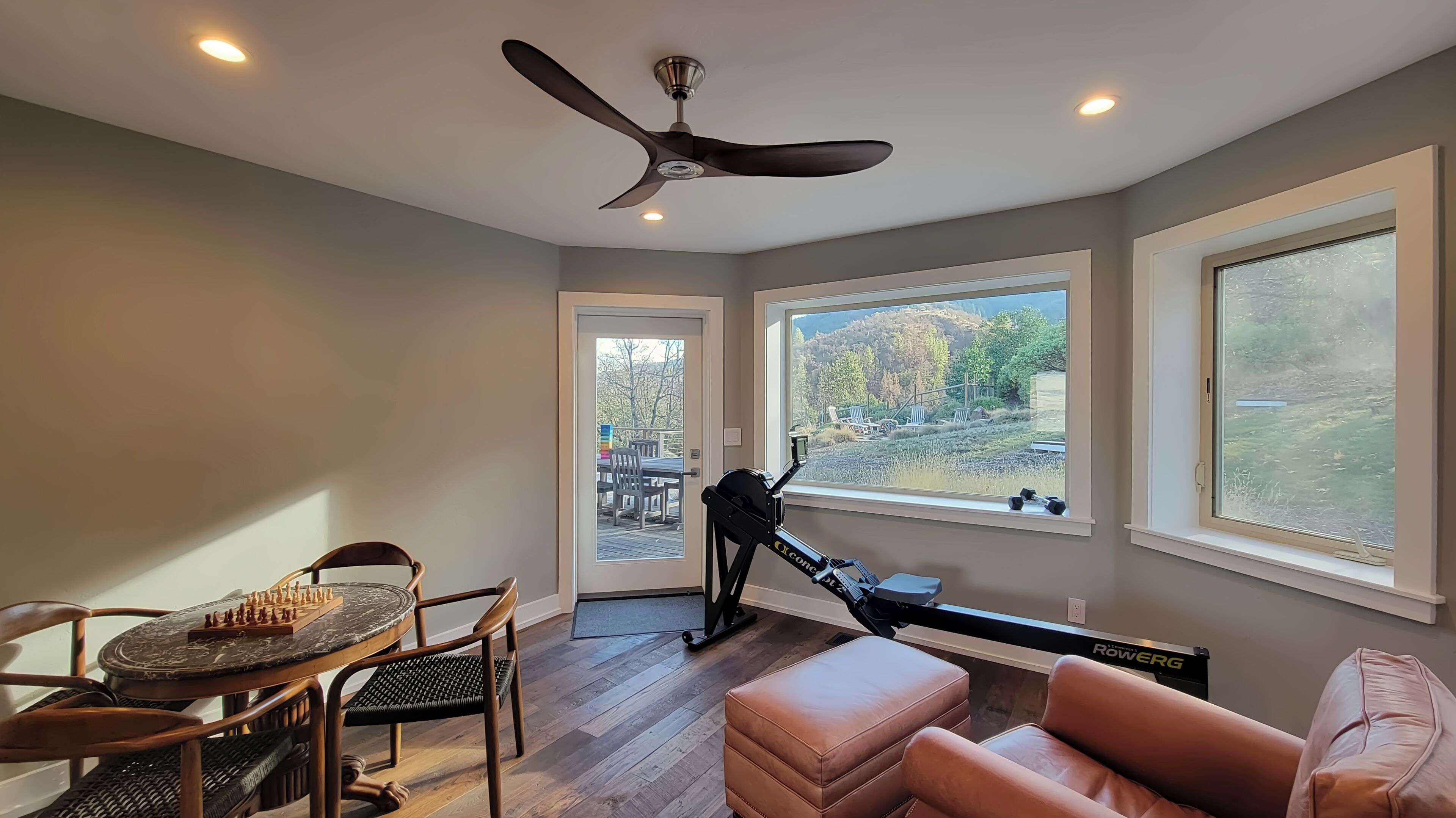 Una habitación con ventilador de techo, cinta de correr, una mesa de comedor con ajedrez y grandes ventanas con vistas al exterior.