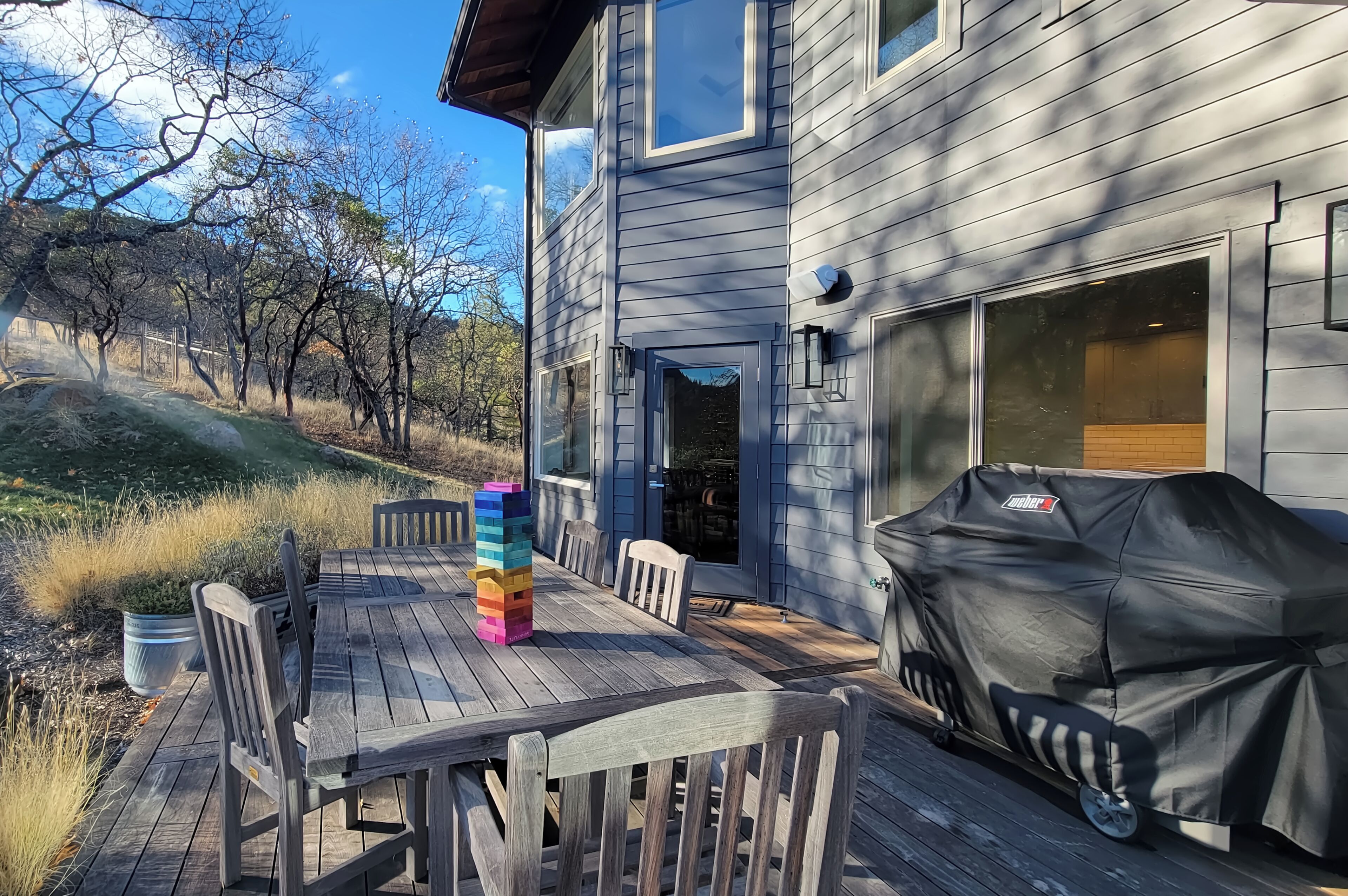 Eine Terrasse mit Holztisch und -stühlen, einem überdachten Grill und Blick auf ein Haus mit Garagentor.