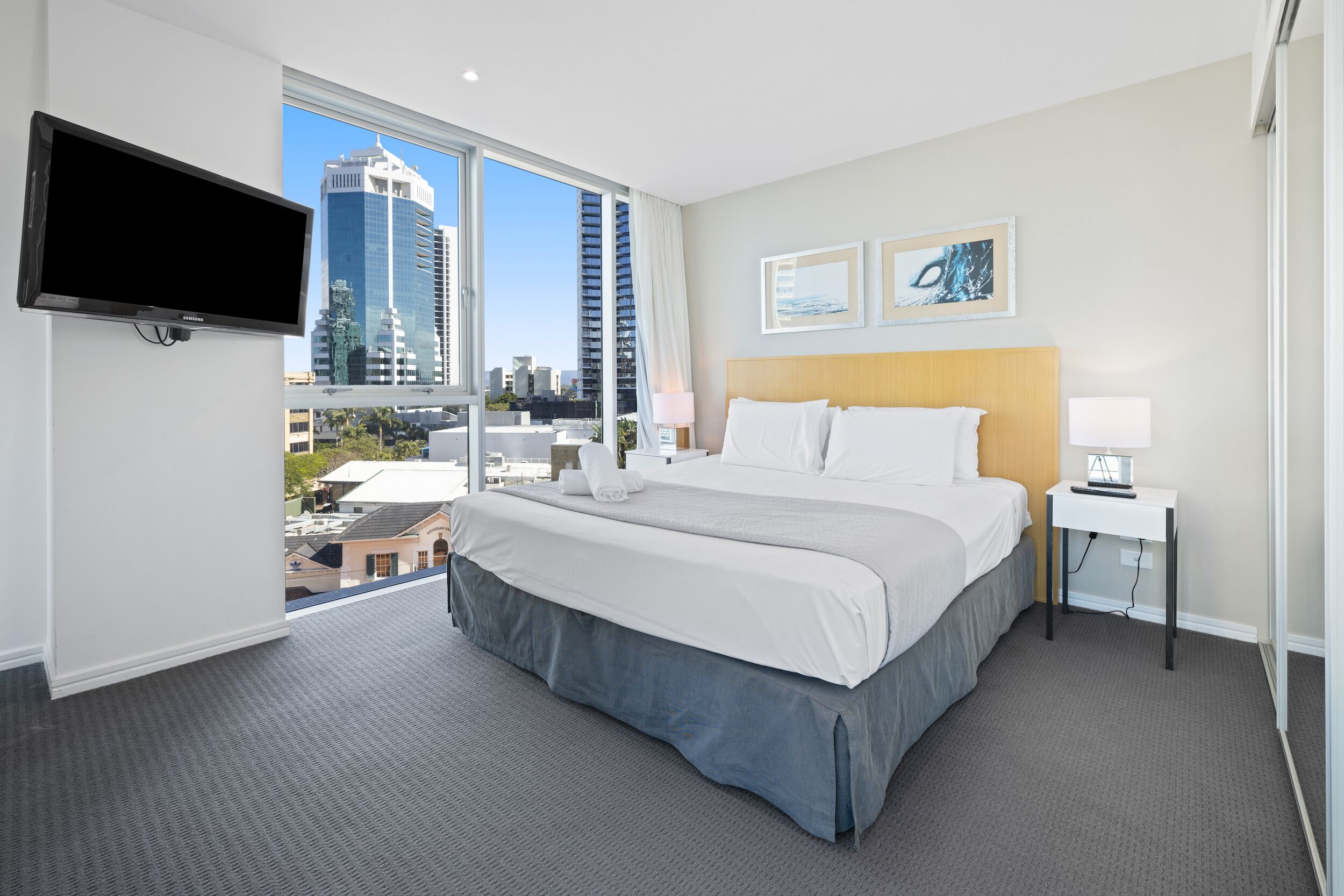 A hotel room with a large bed, a TV mounted on the wall, and a city view through the window.