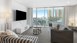 A modern hotel room with a large TV, a grey sofa, a striped ottoman, a small round table, and a balcony with a view of high-rise buildings and the sea. - Pool and Ocean Views at the H-Residences (Surfers Paradise)
