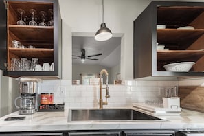 Une cuisine au design moderne, dotée d’un îlot central avec un évier, une machine à café et un pot à café. Des étagères en bois, situées au-dessus de l’îlot, servent à ranger la verrerie, et un ventilateur de plafond est installé au plafond.