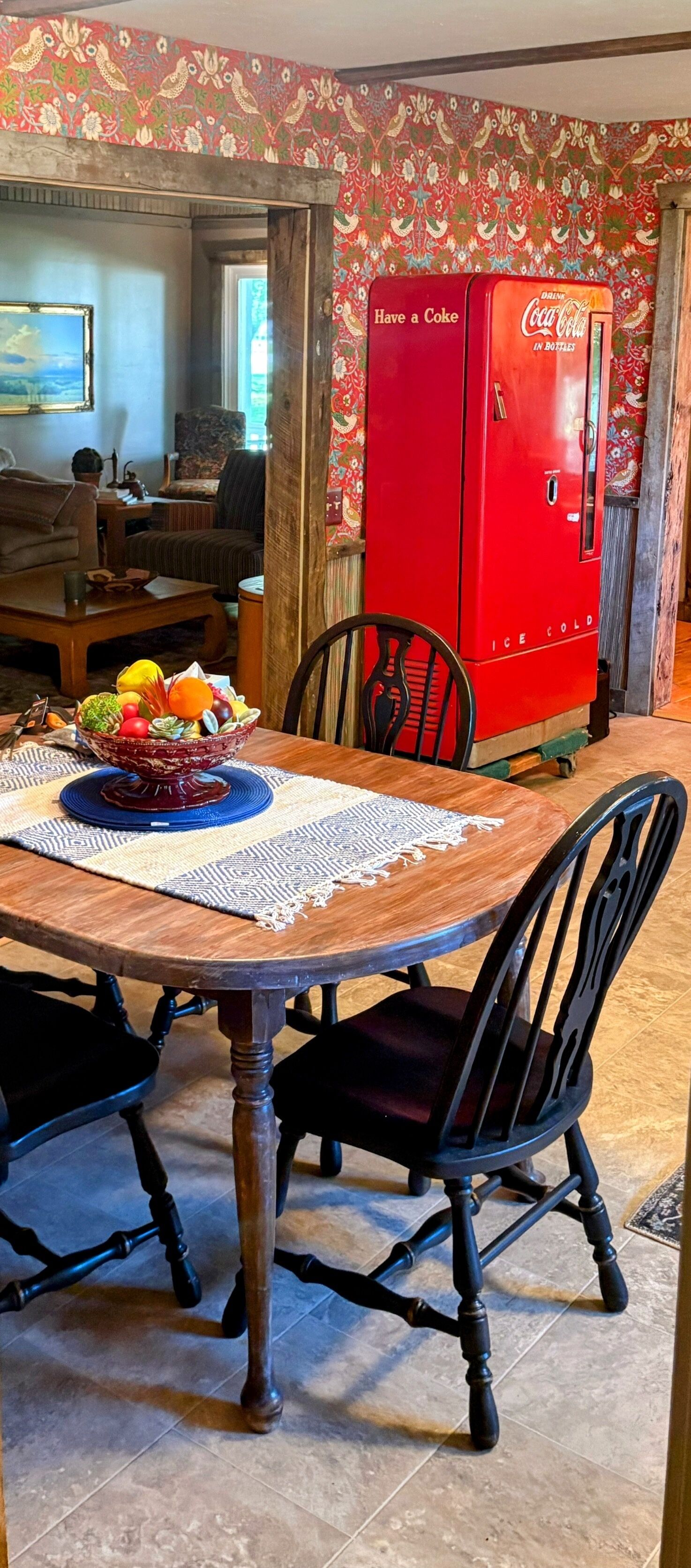 A dining table with a fruit bowl, chairs, and a red Coca-Cola refrigerator.