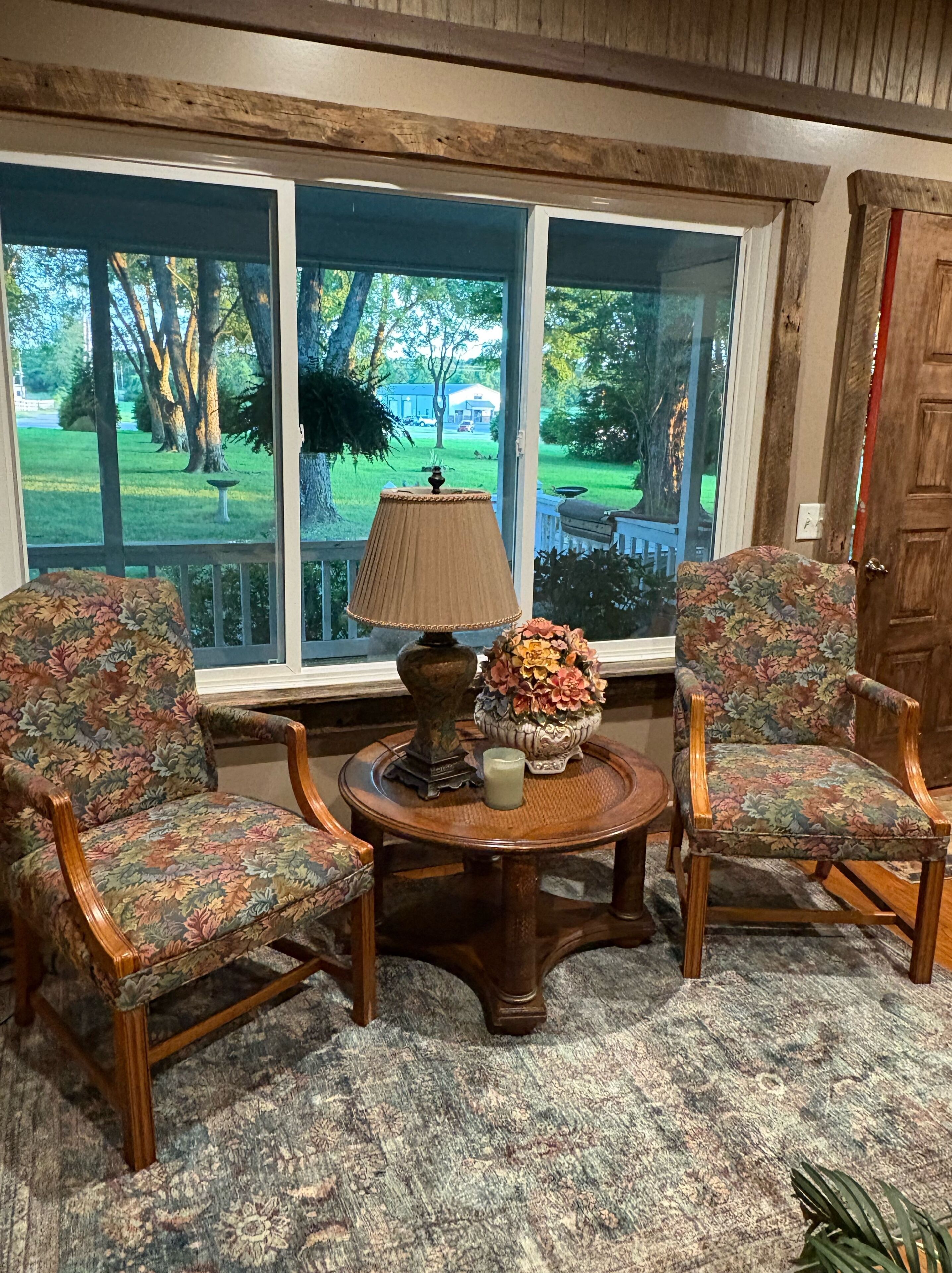 A room with a large window, two floral-patterned chairs, a wooden table with a lamp and vase, and a carpeted floor.