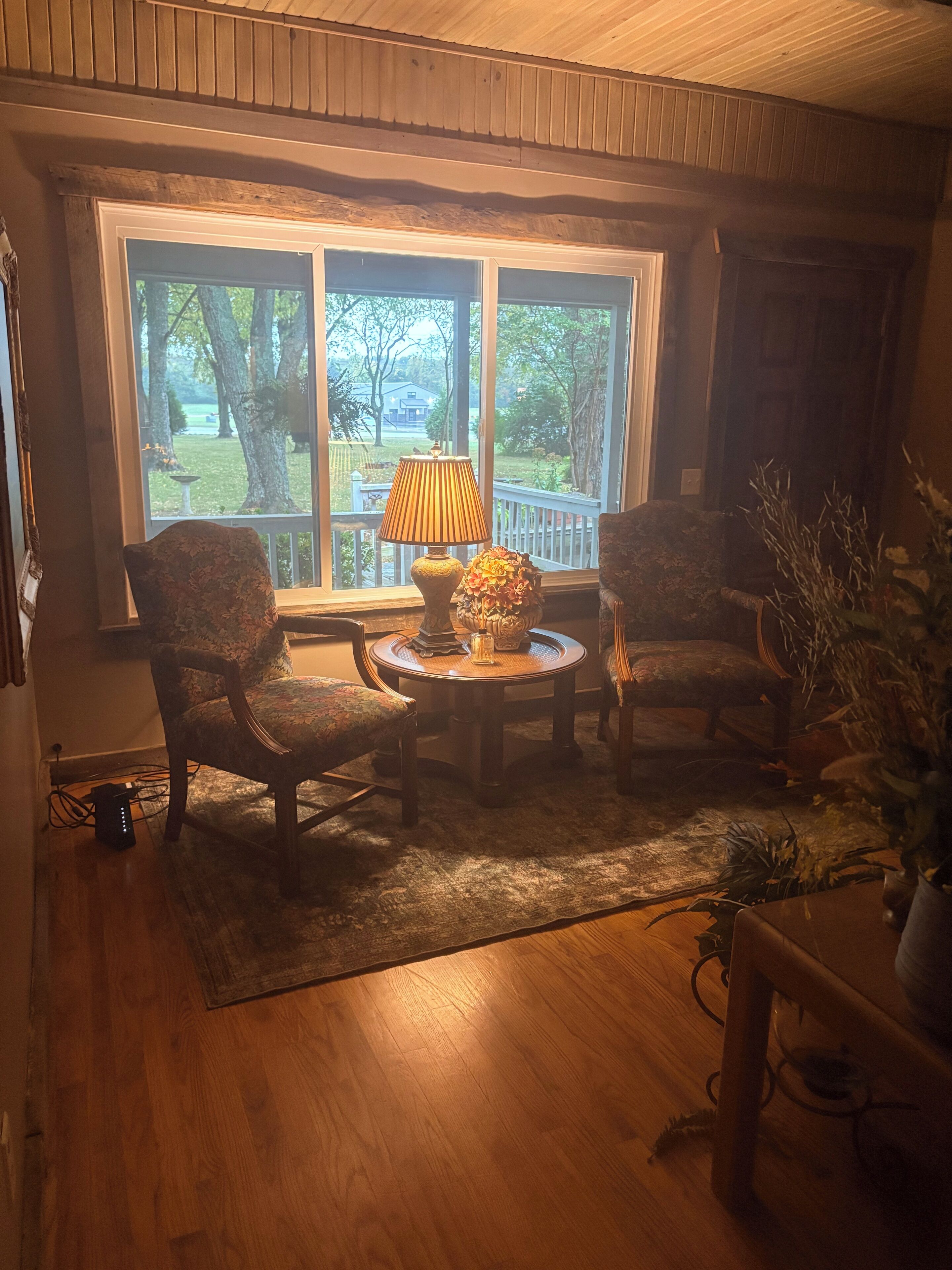 A cozy room with a large window, a round table with a lamp, two chairs, and a potted plant.