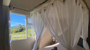 A bedroom with a canopy bed, a window with a view of green hills, and a bedside table with a lamp. - Roucou room in the heart of nature, view of Mont Pelée (Le Morne Rouge)