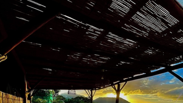 A wooden deck with outdoor furniture, a hammock, and a view of a sunset over a landscape. - Roucou room in the heart of nature, view of Mont Pelée (Le Morne Rouge)
