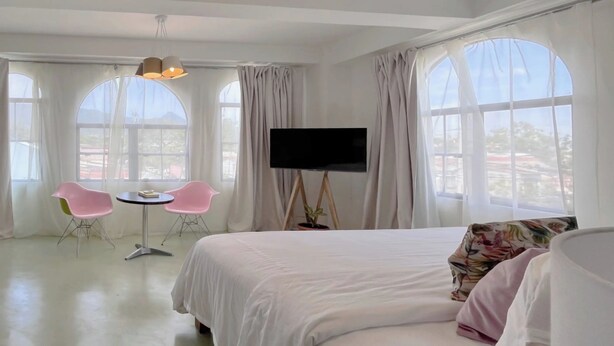 A hotel room with a bed, a TV on a stand, two pink chairs, and large windows with curtains. - Ipan Noya (Santa Tecla)