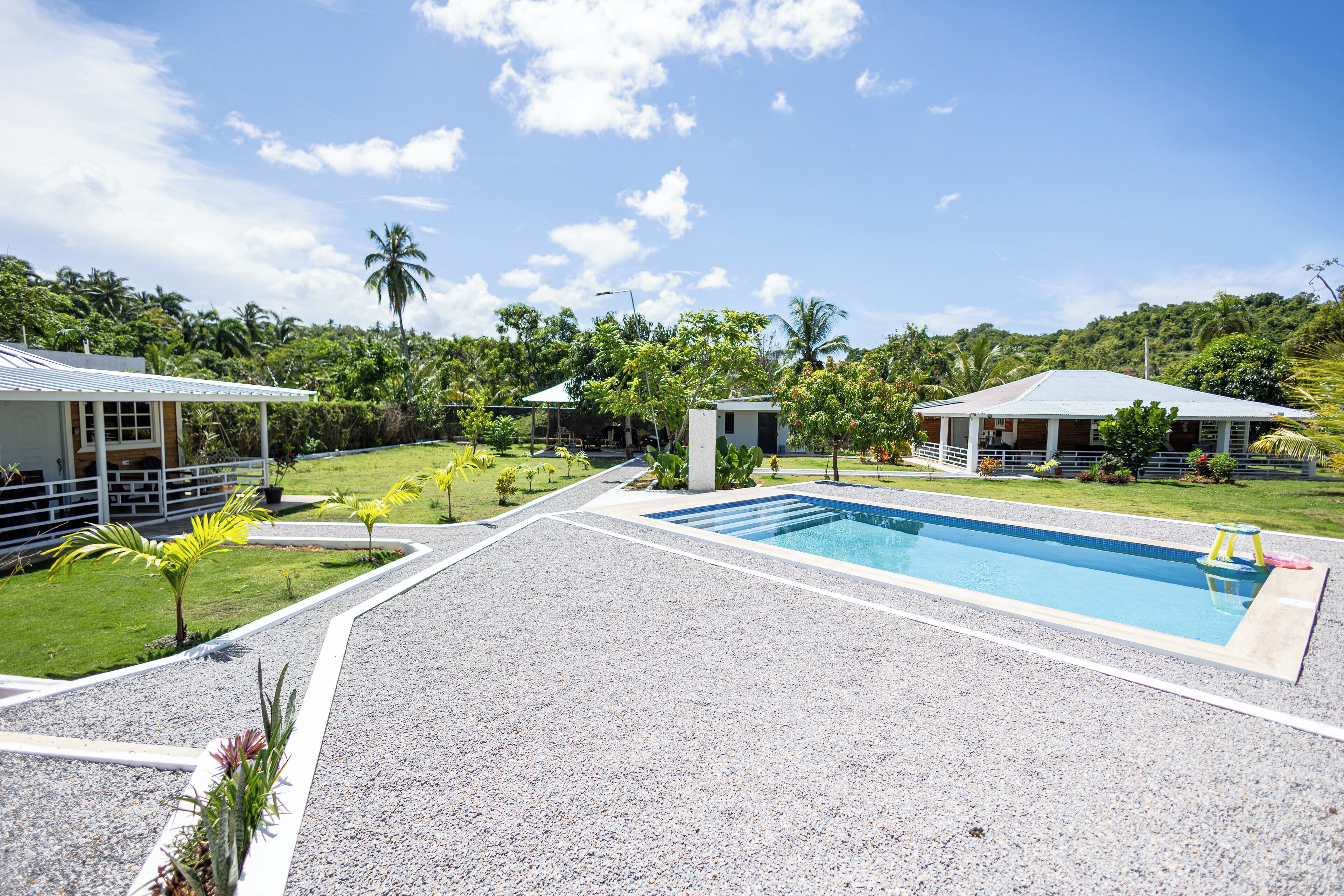 A swimming pool surrounded by a gravel area, with a small building and a covered structure nearby.
